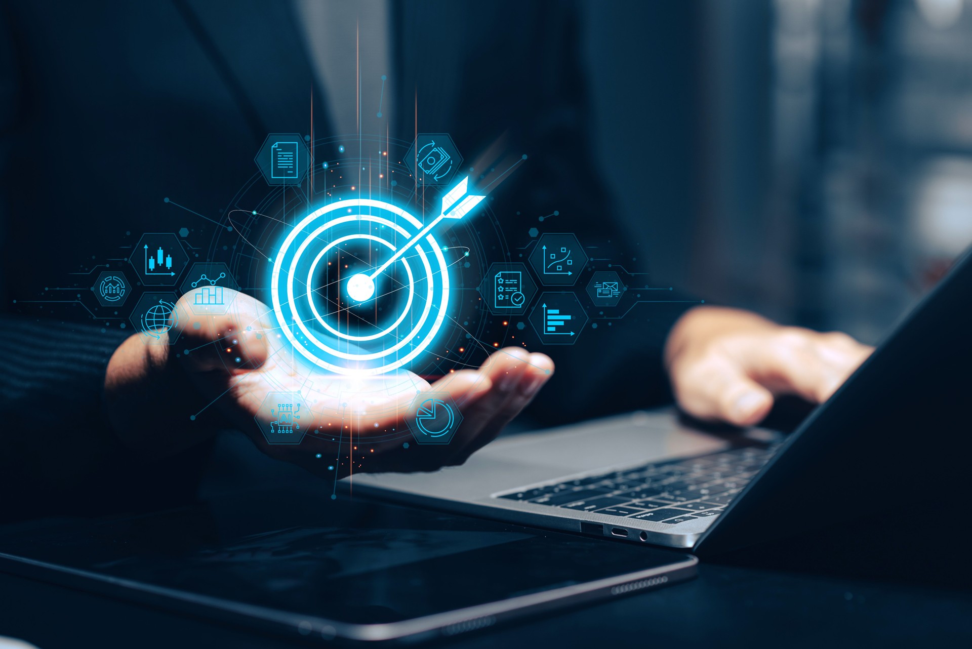 Businessman holding a glowing blue target with arrow in his palm above a laptop, surrounded by holographic business icons representing goals, analytics, strategy, and online presence. This conceptual image embodies business target achievement, success.